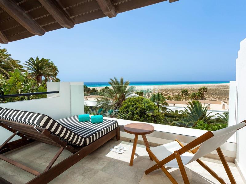 Balcón con tumbona moderna y silla con vista al mar y palmeras.