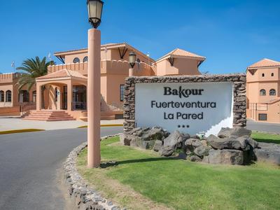Entrance area with a large sign in front of a Mediterranean-style hotel complex under a blue sky.