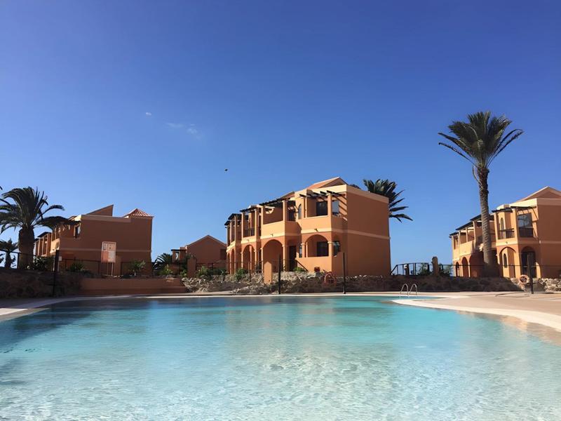 View of a large pool and orange hotel buildings under clear blue sky with palm trees.