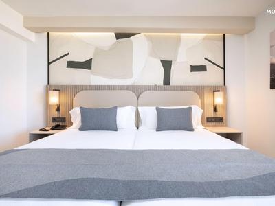 Camera d'hotel moderna con letto matrimoniale, copriletto grigio e testiera con arte astratta.