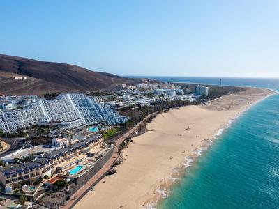 Vista aerea di una spiaggia con hotel e colline sullo sfondo sotto un cielo limpido.