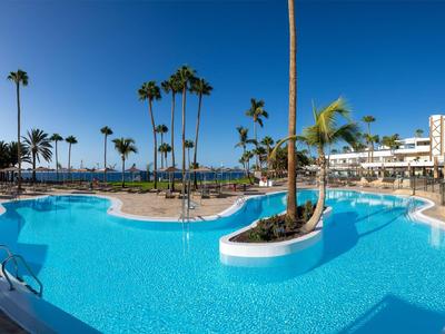 Grande piscina all'aperto con palme e edifici dell'hotel sotto un cielo azzurro limpido.