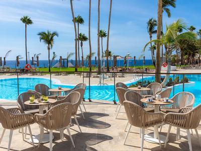 Stuhl- und Tischgruppe am hotelpool mit Palmen und Meerblick unter blauem Himmel.