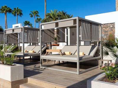 Lounge esterna moderna con aree coperte e cuscini accanto a piante tropicali.