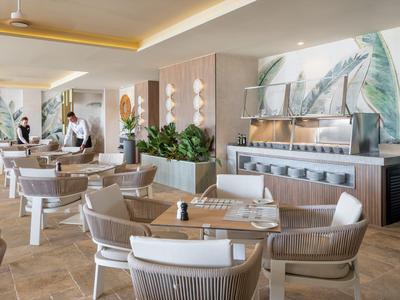 Ristorante luminoso e moderno con gruppi di tavoli, sedie e piante decorative.