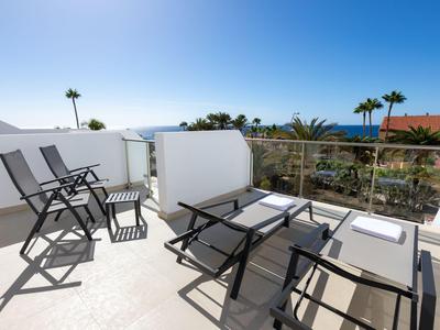 Terrasse mit Liegestühlen und Stühlen, Blick auf Palmen und Meer bei klarem Himmel.