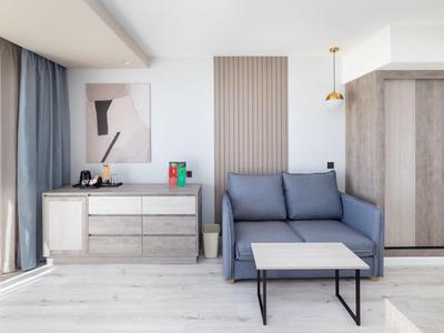 Modern eingerichtetes Hotelzimmer mit Sofabett, Kommode und großem Fenster mit Vorhängen.