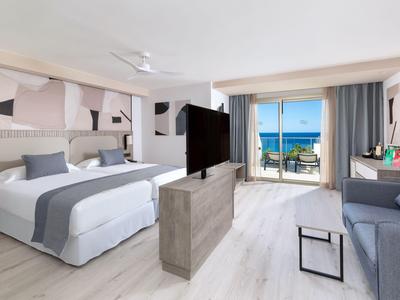 Helles Hotelzimmer mit Doppelbett, Sofa, TV und Balkon mit Meerblick.