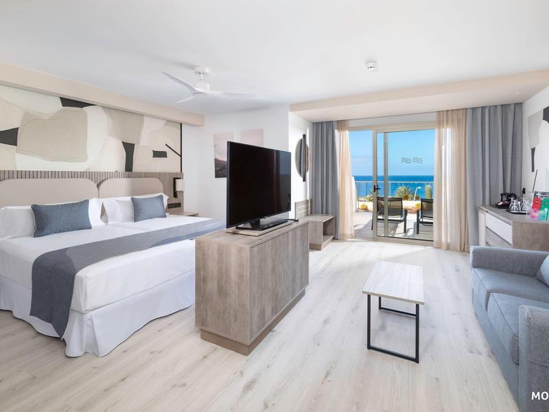 Camera d'hotel moderna con letto matrimoniale, TV, divano e vista mare dal balcone.