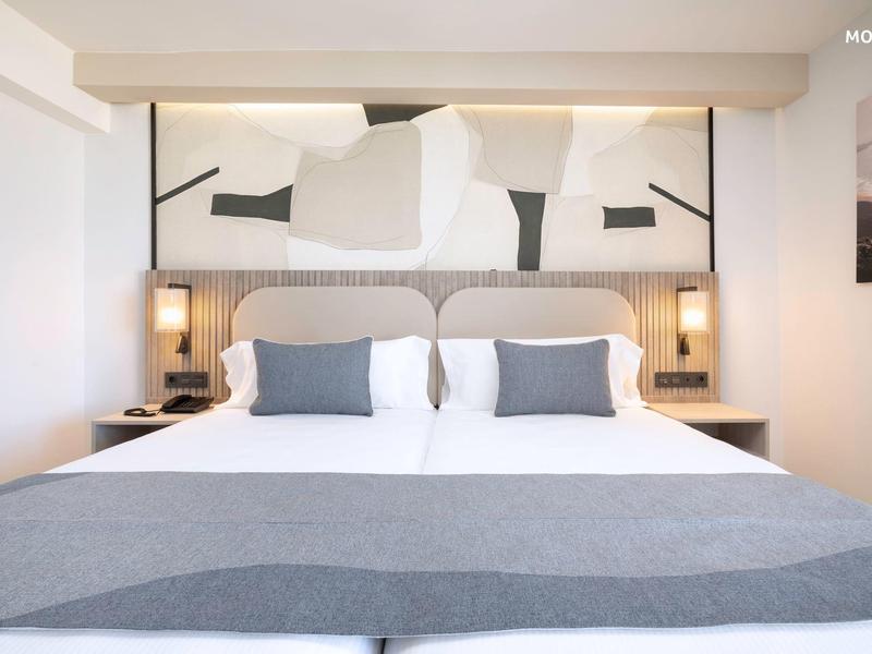 Camera d'hotel moderna con letto matrimoniale, copriletto grigio e testiera con arte astratta.