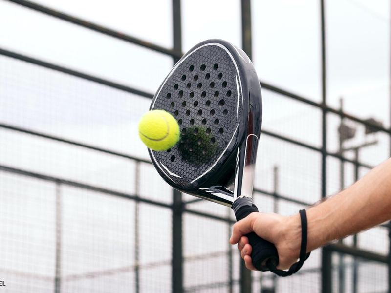 Primo piano di una racchetta da padel che colpisce una pallina da tennis davanti a una recinzione.