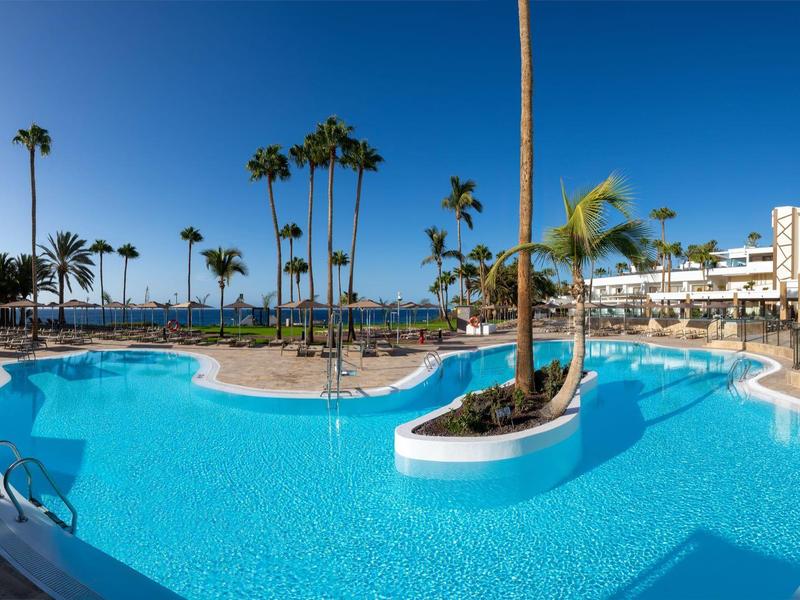 Grande piscina all'aperto con palme e edifici dell'hotel sotto un cielo azzurro limpido.