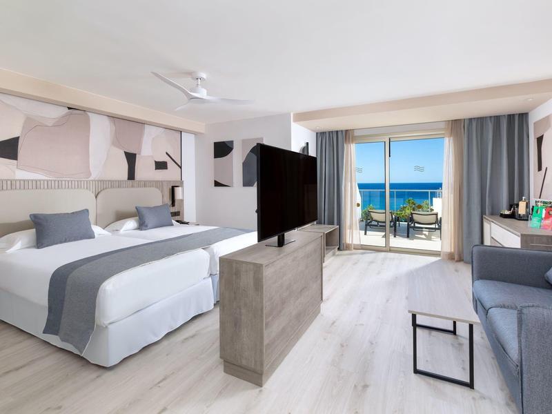 Camera d'hotel luminosa con letto matrimoniale, divano, TV e vista mare dal balcone.