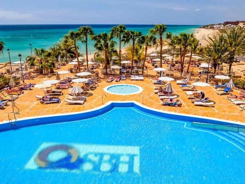 Grande piscina dell'hotel con lettini e palme vicino alla spiaggia sotto un cielo limpido.