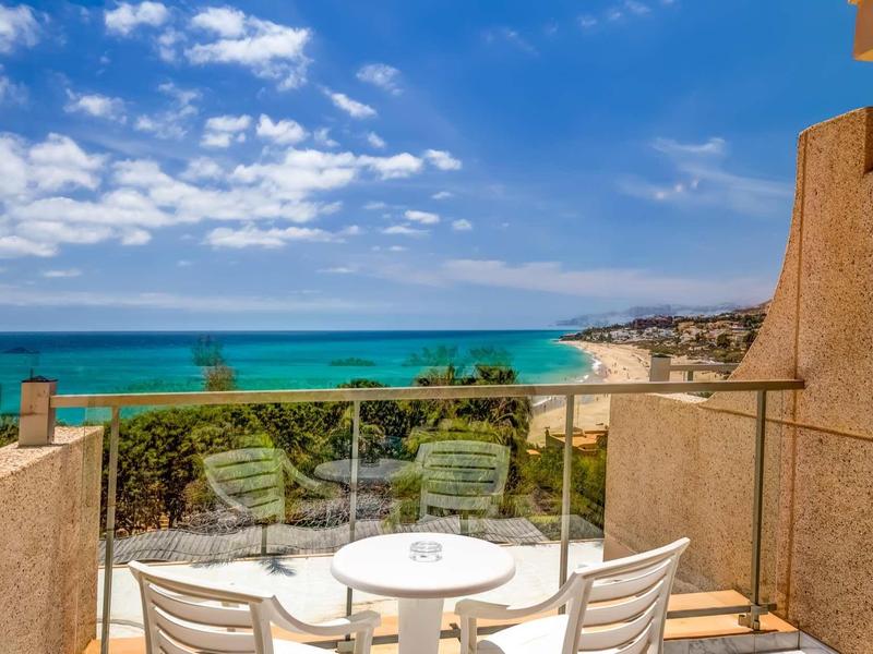 Balcone con tavolo rotondo e sedie con vista sul mare blu e spiaggia sabbiosa sotto un cielo azzurro.