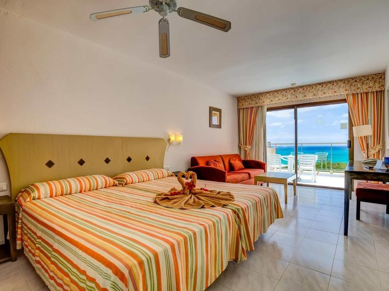 Camera d'hotel luminosa con letto matrimoniale, ventilatore a soffitto, divano e vista mare dal balcone.