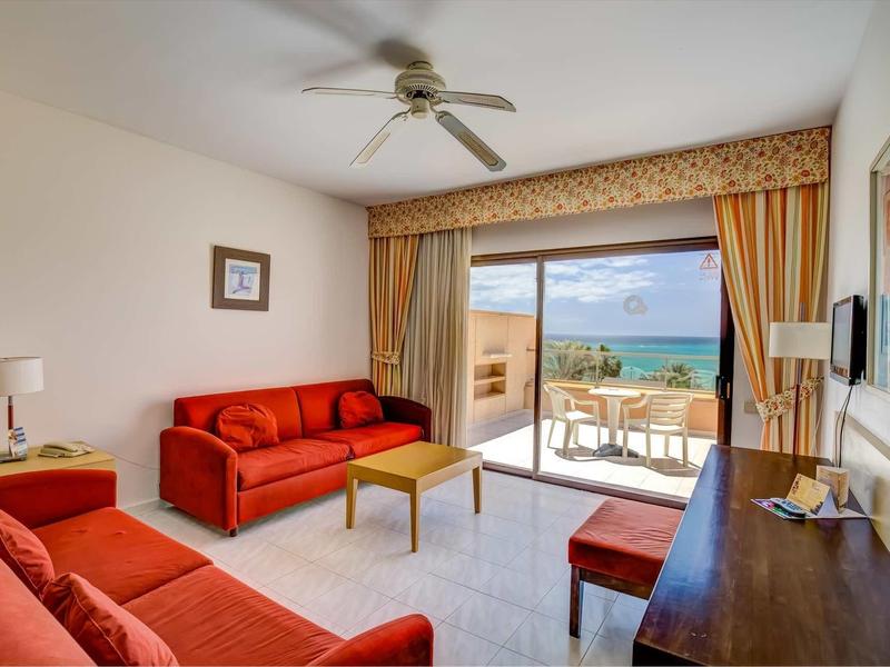 Soggiorno luminoso con divani rossi, ventilatore a soffitto e vista mare dalla grande porta del balcone.