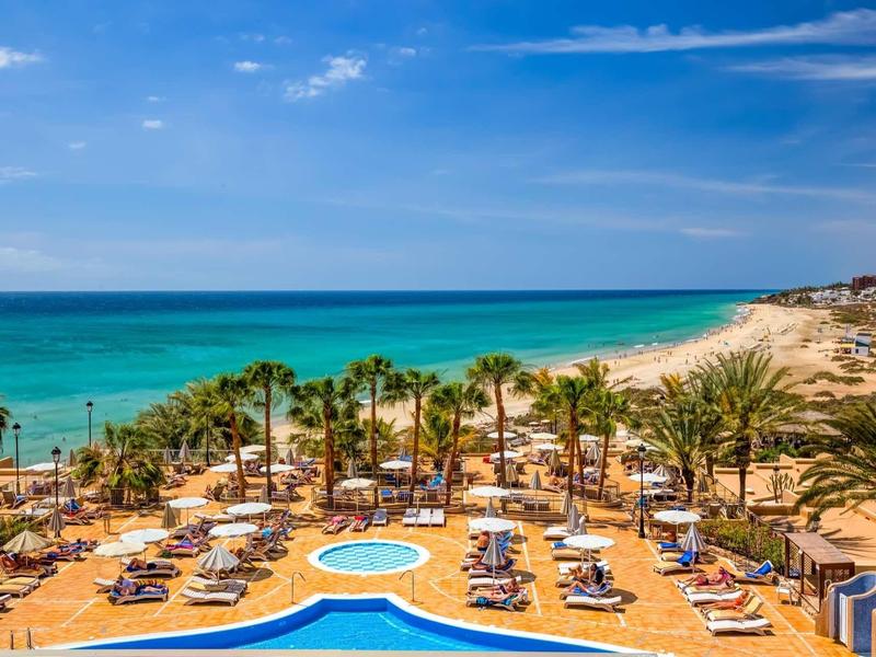 Resort sulla spiaggia con piscina, lettini e palme sotto il cielo azzurro vicino al mare turchese.