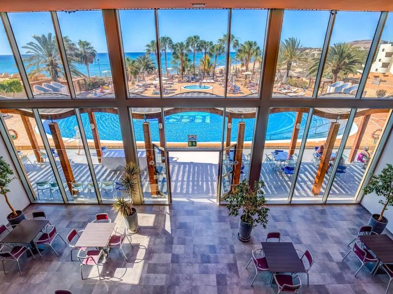 Vista attraverso grandi finestre di una piscina e palme da una zona pranzo interna.