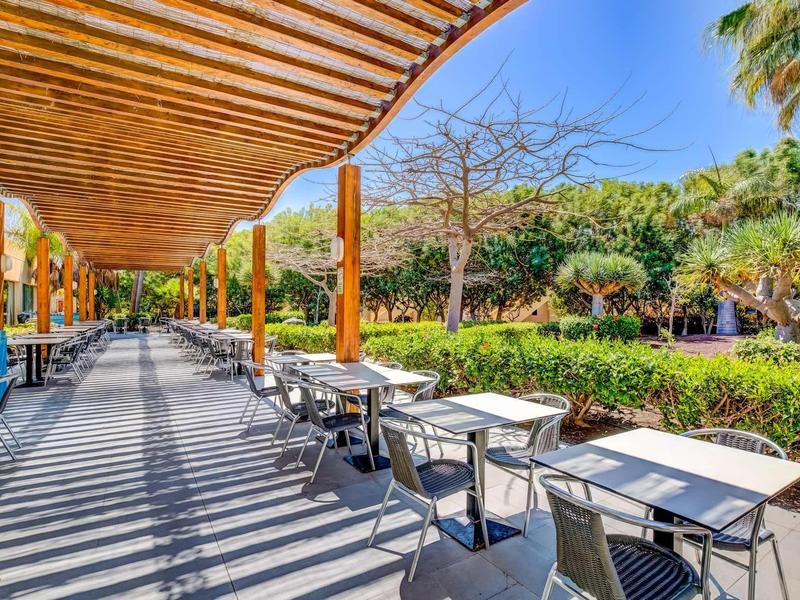 Area esterna soleggiata con tavoli e sedie sotto una pergola nel giardino di un hotel.