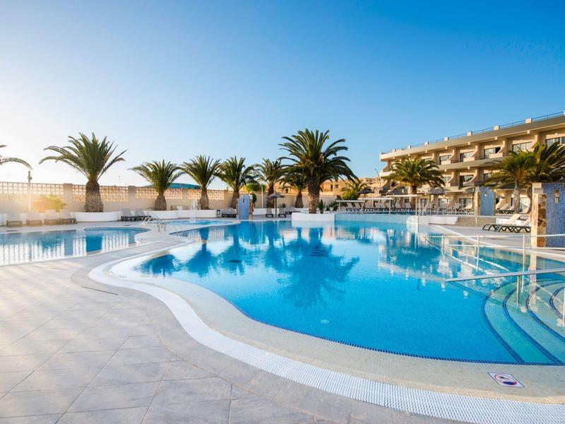 Piscina esterna dell'hotel con palme e lettini sotto un cielo azzurro limpido al tramonto.