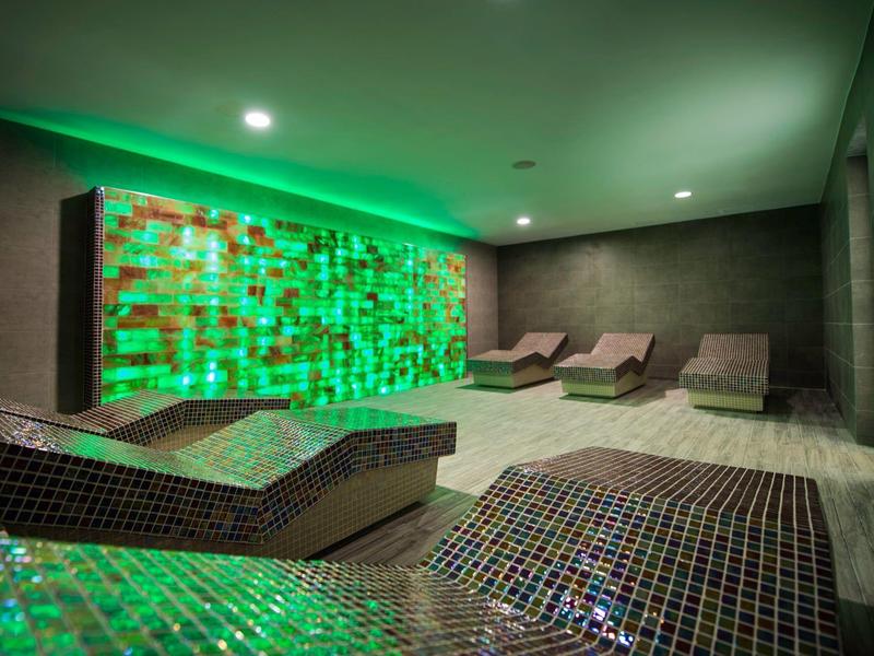 Spa moderna con chaise longue a mosaico e parete di vetro verde illuminata sotto faretti incassati.
