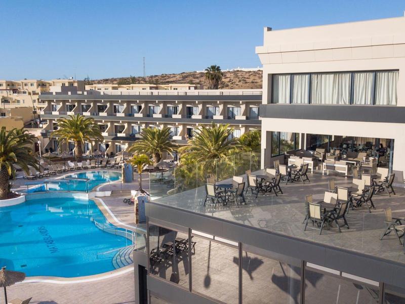 Hotel con grande piscina all'aperto, palme e area ristorazione sotto un cielo azzurro.