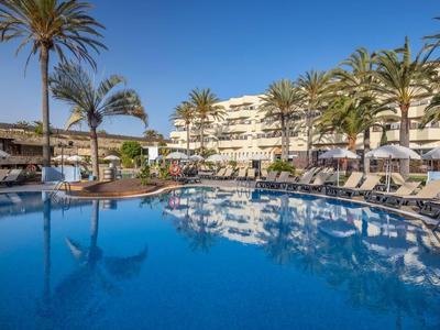 Sunny hotel pool surrounded by lounge chairs and palm trees under clear blue sky.