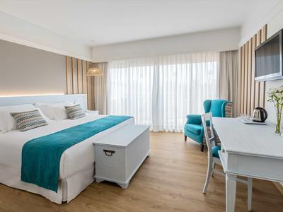 Bright hotel room with large bed, teal accents, desk, armchair and sheer curtains over balcony door.