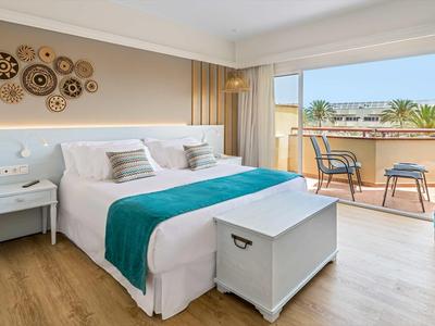 Bright hotel room with large bed, balcony, turquoise accents, and wooden floor.