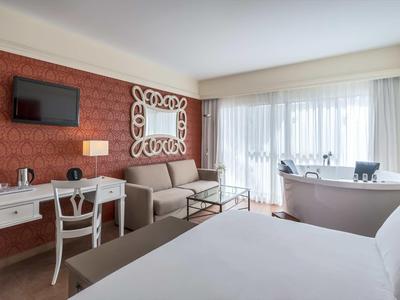 Bright hotel room with bed, sofa, desk, TV, and large window with sheer curtains.