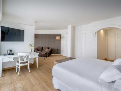 Bright hotel room with large bed, seating area, desk, and wall-mounted TV.