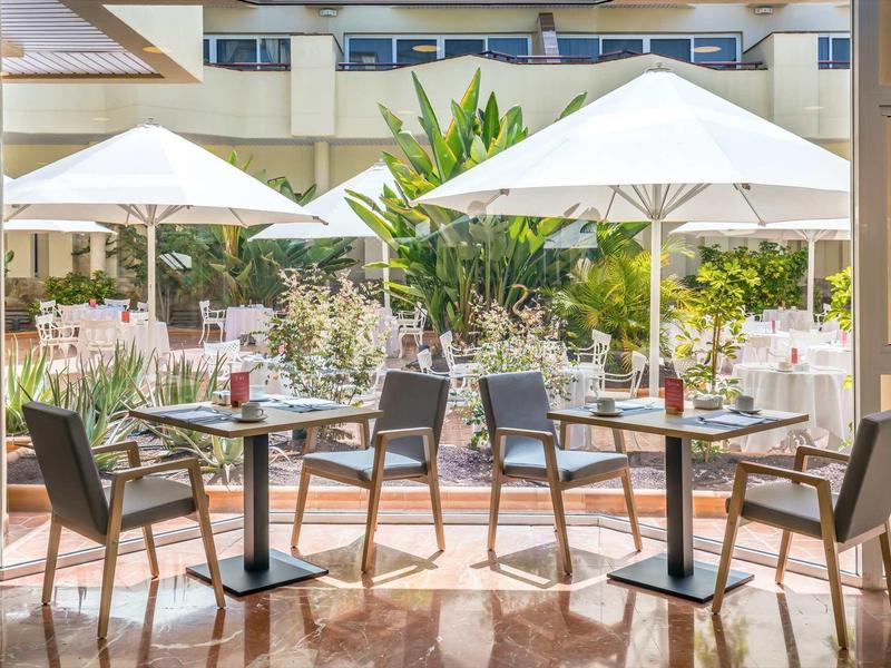 Outdoor dining area with tables, chairs, umbrellas, and lush greenery in a hotel courtyard.