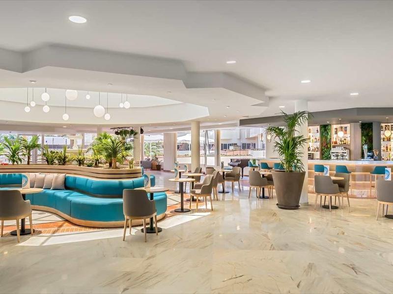 Spacious hotel lobby with modern blue seating, wooden furniture, and large potted plants under bright lighting.