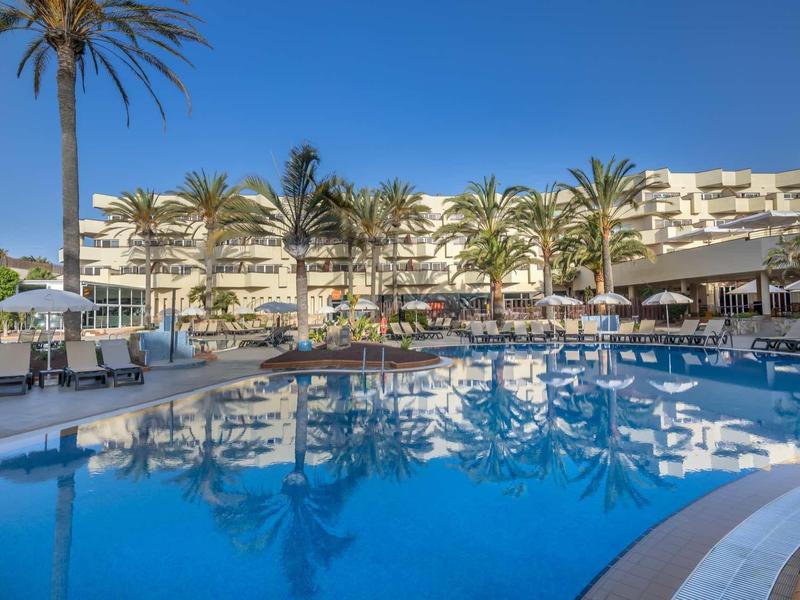 Large hotel pool surrounded by palm trees, lounge chairs, and white buildings under clear sky.