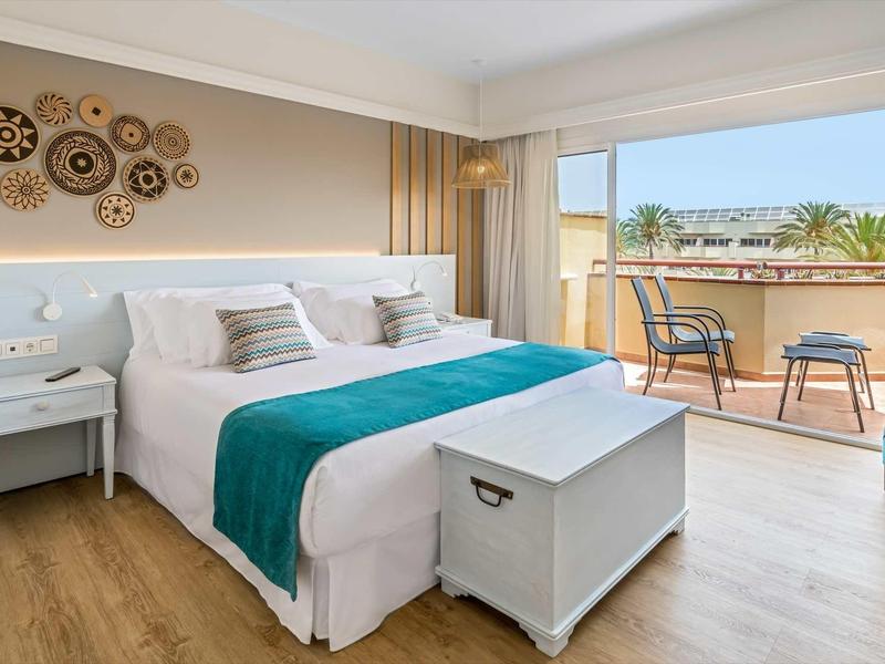 Bright hotel room with large bed, balcony, turquoise accents, and wooden floor.