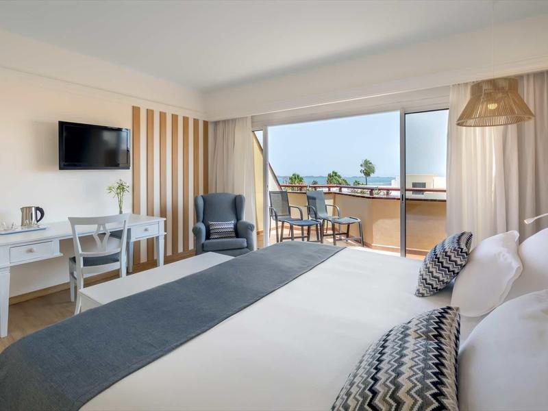 Bright hotel room with large bed, balcony with chairs, desk, and TV mounted on the wall.