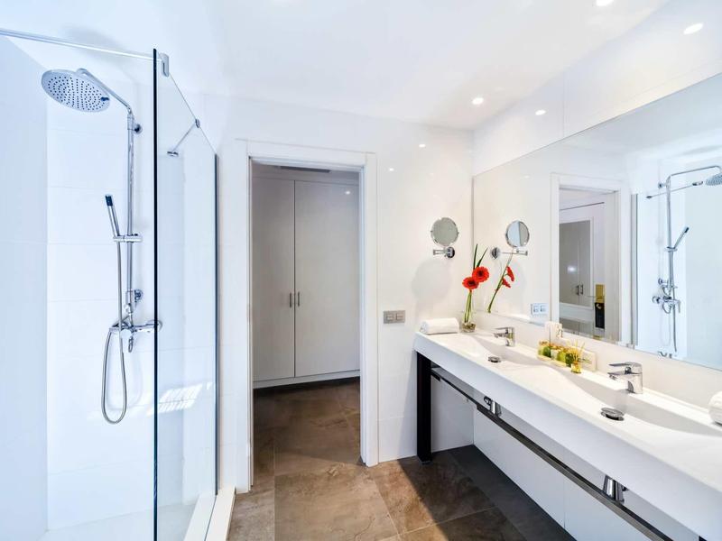 Bright modern bathroom with shower, long mirror, double sinks, and brown tiled floor.