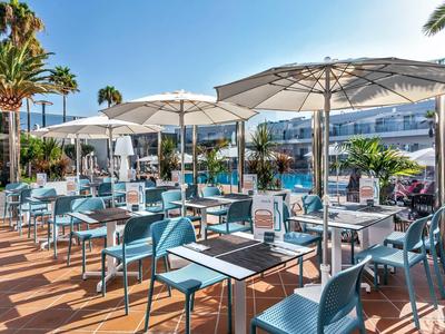 Sunny outdoor hotel terrace with blue chairs, tables, and large white umbrellas by a pool.