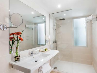 Modern white bathroom with double sink, large mirror, glass shower, and a vase with red flowers.