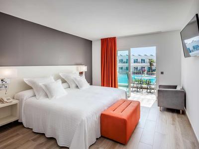 Bright hotel room with a large bed, orange bench, and balcony with pool view.