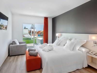 Bright hotel room with a large bed, TV, armchair, and glass door leading to a garden patio.