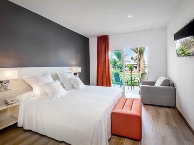 Modern hotel room with a large bed, sofa, TV, and balcony overlooking greenery.