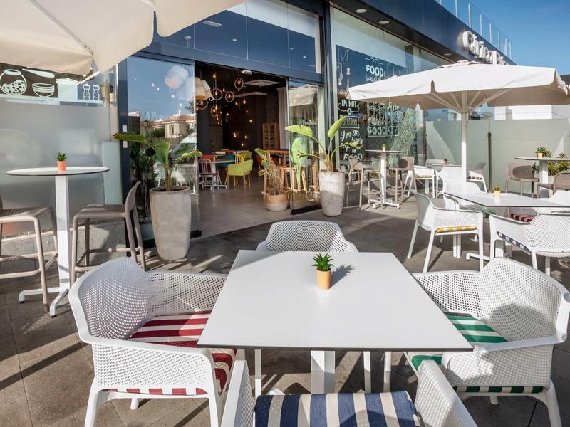 Outdoor restaurant seating with white tables, chairs, umbrellas, and open entrance to interior.