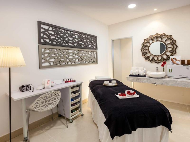 Modern spa room with massage table, decorative mirror, and a desk with chair and lamp.
