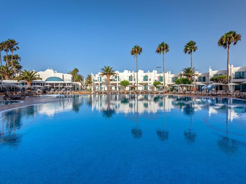 Large outdoor pool with palm trees and white buildings under a clear blue sky.