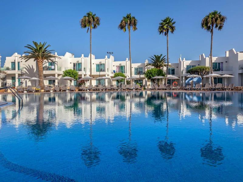 Resort with palm trees and white buildings reflected in a large blue swimming pool.