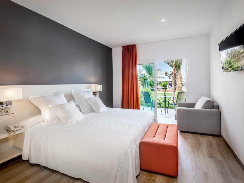 Modern hotel room with a large bed, sofa, TV, and balcony overlooking greenery.