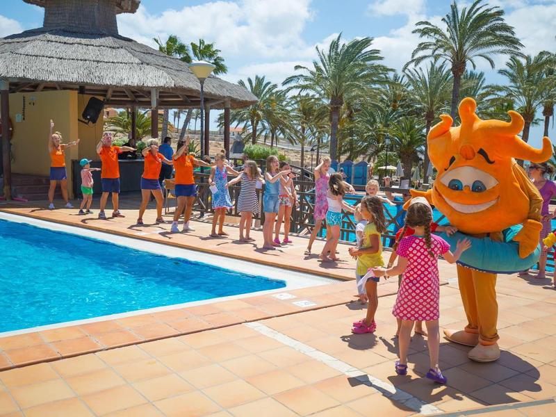 Bambini che corrono vicino alla piscina accanto a un bar dell'hotel, accompagnati da una mascotte arancione.