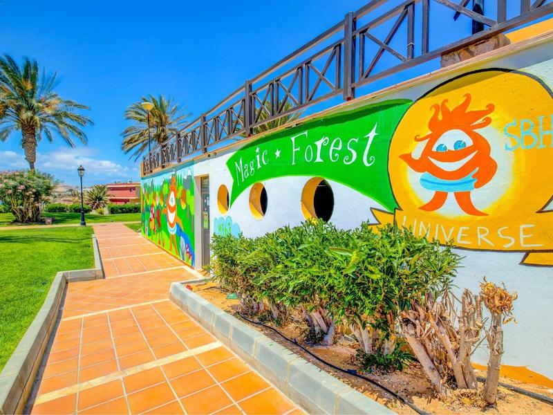 Muro dipinto colorato con buchi davanti a un giardino curato e palme sotto un cielo azzurro.